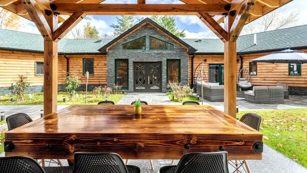 Una cabaña de madera con entrada de piedra, un porche cubierto con mesa y sillas, y un jardín con mobiliario exterior.