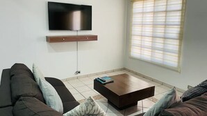 A living room with a flat-screen TV mounted on the wall, a wooden coffee table, and a large window with blinds. - 3 Bedrooms Home 5 min Walking Distance to El Malecon (La Paz)