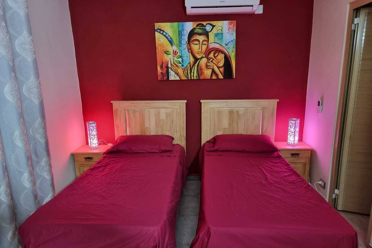 Ein Zimmer mit zwei Betten, jeweils mit einem hölzernen Kopfende und einer roten Bettdecke. An beiden Betten befinden sich Nachttische mit dekorativen Lampen, darüber ein Gemälde.