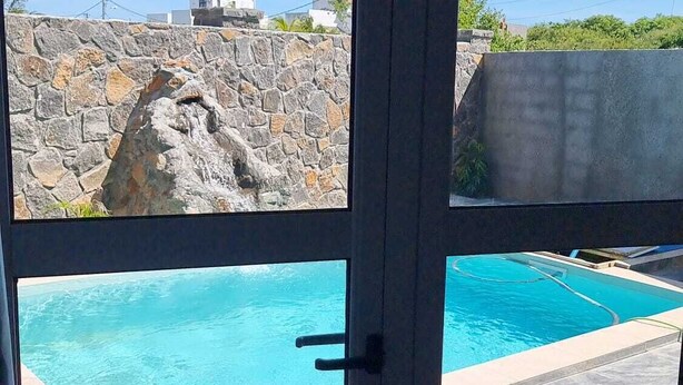 A view through a glass door to a swimming pool with a stone wall and a clear blue sky. - Doux Réveil (Cap Malheureux)