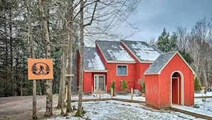 Una casa roja con una construcción pequeña adosada, rodeada de árboles y suelo cubierto de nieve.
