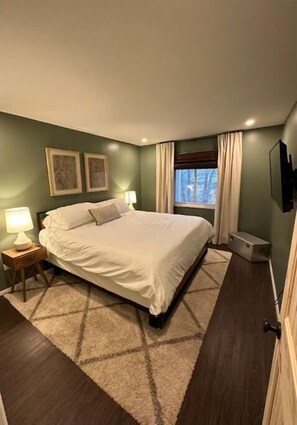 Een slaapkamer met een groot bed, twee bedlampen, een raam met gordijnen en een flatscreen televisie aan de muur.