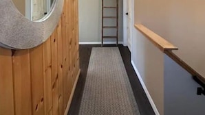 Un pasillo con pared de madera, un espejo circular, un felpudo con motivo de espiga, una escalera de madera y una puerta con un ventanal encima.
