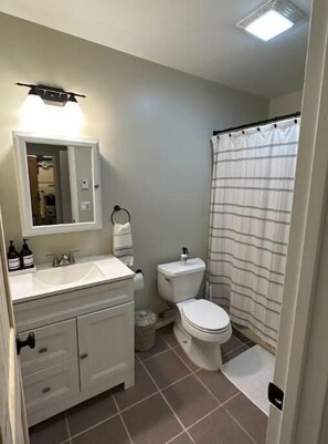 Een badkamer met een witte wastafel, spiegel en toilet. Er is een douchegordijn en een kleine prullenbak.