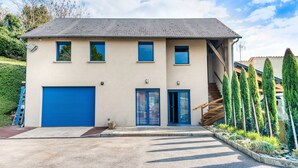A two-story building with a blue garage door, a staircase leading to the upper floor, and a row of tall, narrow trees in front. - Gîte du Thouet - Appt avec piscine partagée (Montreuil-Bellay)