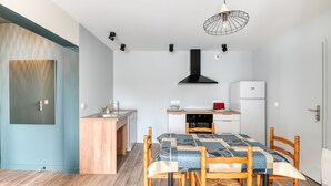 A compact kitchen with a dining area, wooden chairs, a table with a patterned tablecloth, a black range hood, and a white refrigerator. - Gîte du Thouet - Appt avec piscine partagée (Montreuil-Bellay)