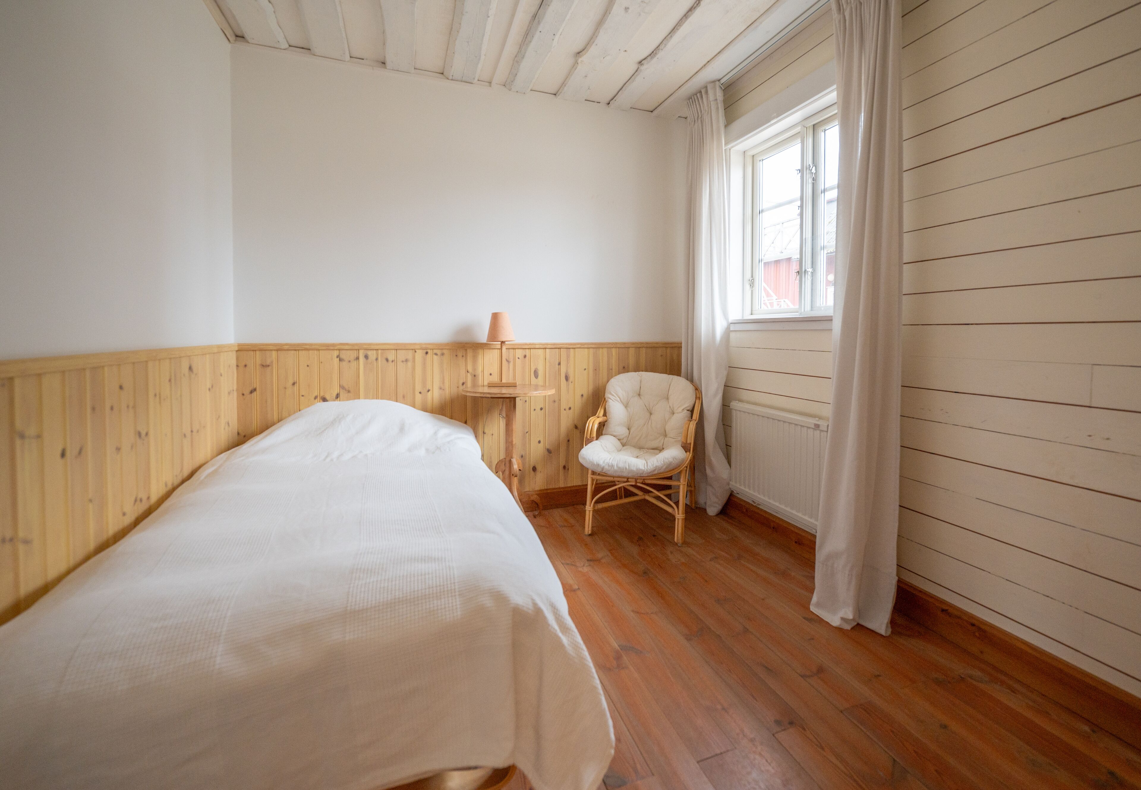 A room with a single bed, a chair, a window with curtains, and wooden walls.