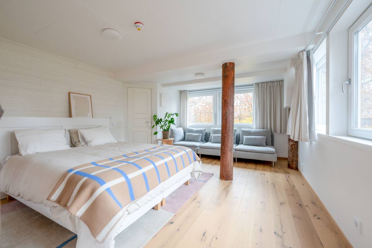 A modern bedroom with a large bed, a wooden column, a sofa, and a window with curtains.