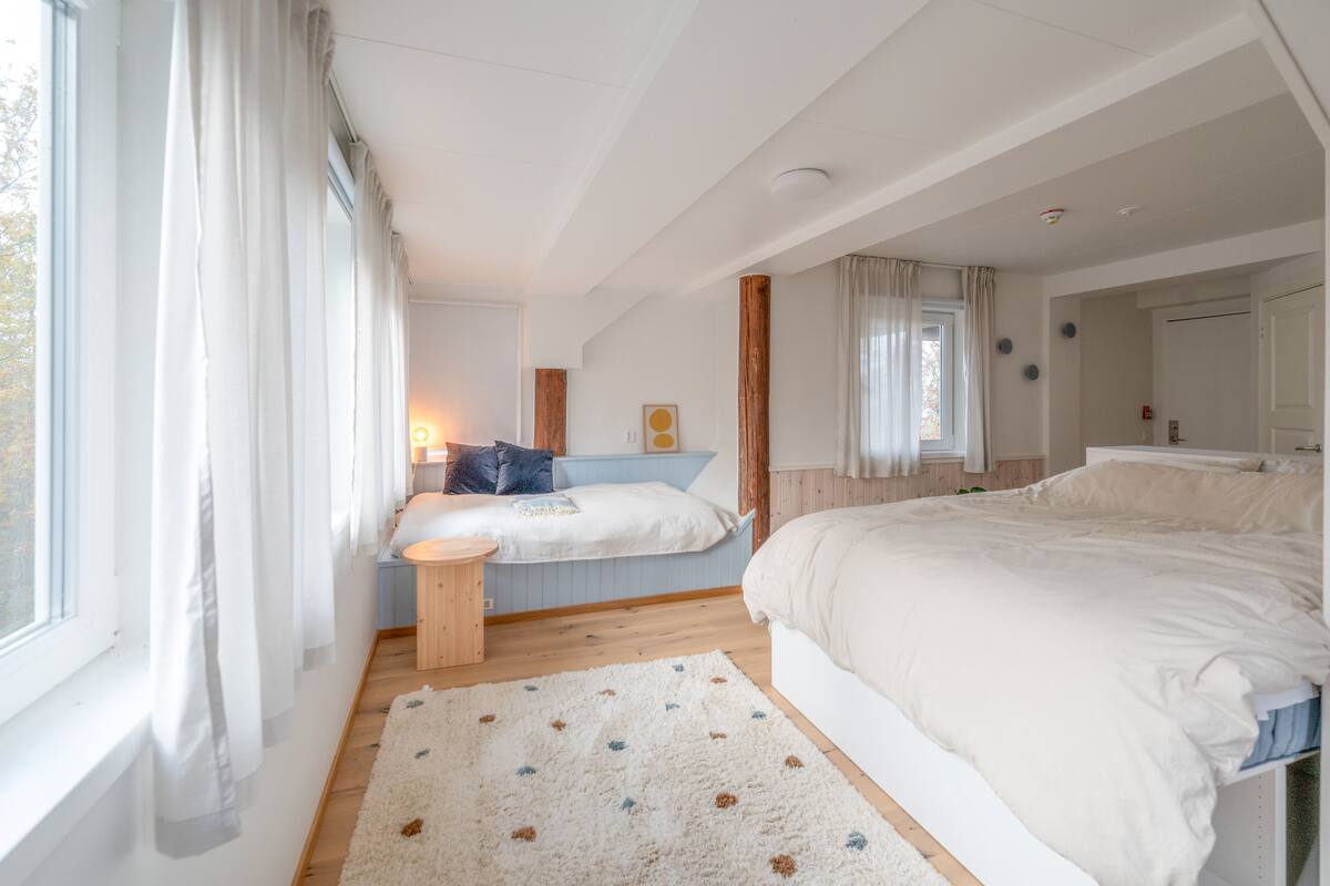 A bedroom with two beds, a large window with curtains, a wooden floor, and a small round table.