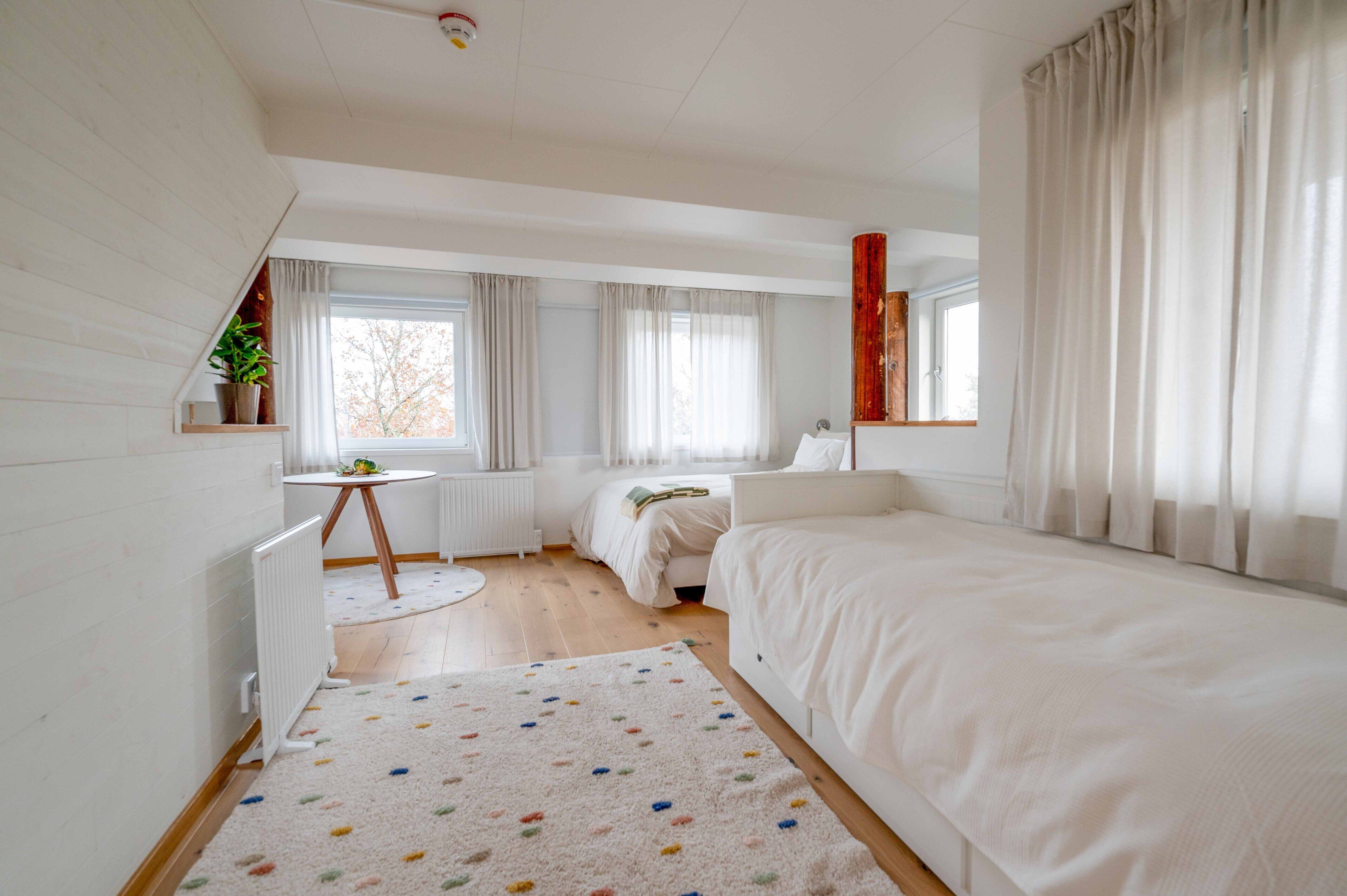 Un dormitorio moderno con una cama grande, una cama más pequeña, una mesa redonda con una planta y una ventana con cortinas.