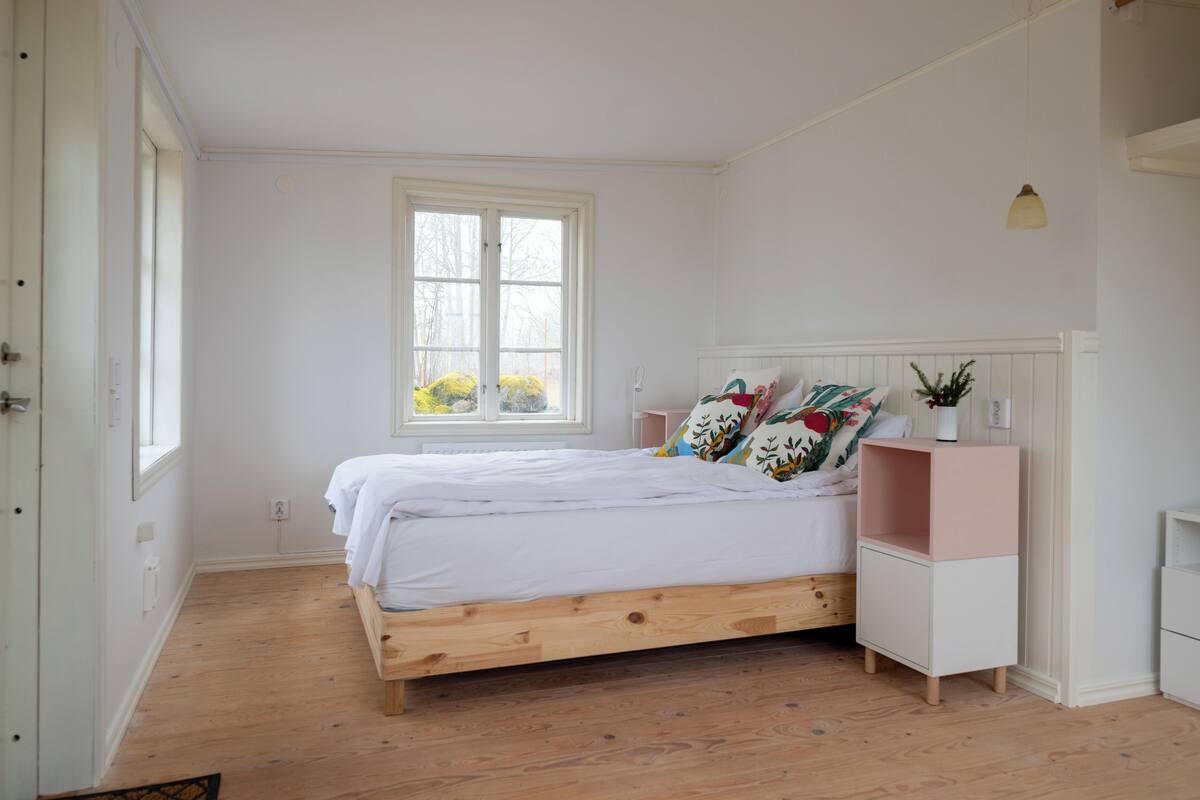 A bedroom with a wooden bed, white bedding, a bedside table, and a window with a view of greenery.
