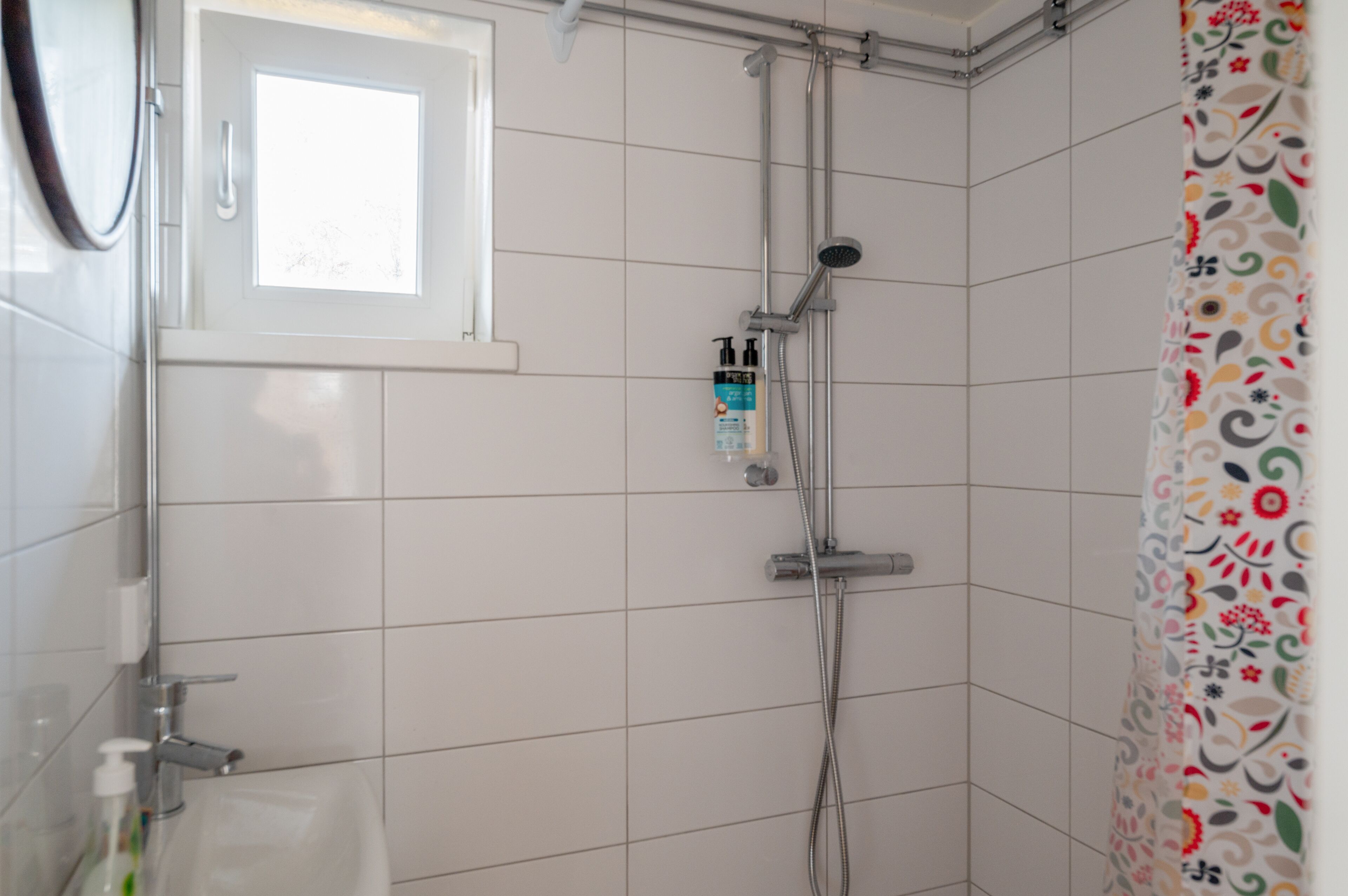 Une salle de bain avec des murs recouverts de carreaux blancs, une douche avec un pommeau de douche amovible, une fenêtre et un rideau de douche coloré.