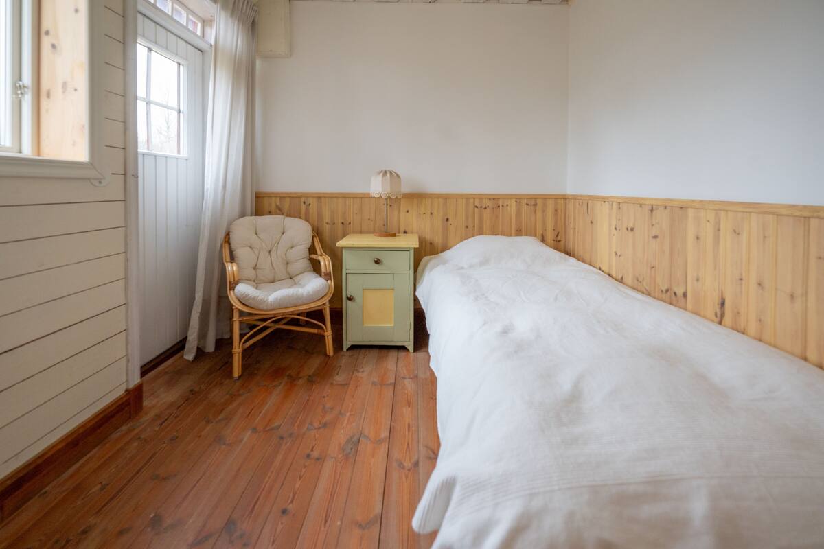 A room with a wooden floor, a bed, a chair, a nightstand, and a window with curtains.