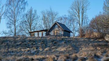 A wooden cabin with a sloped roof, situated on a grassy hill with leafless trees in the background.