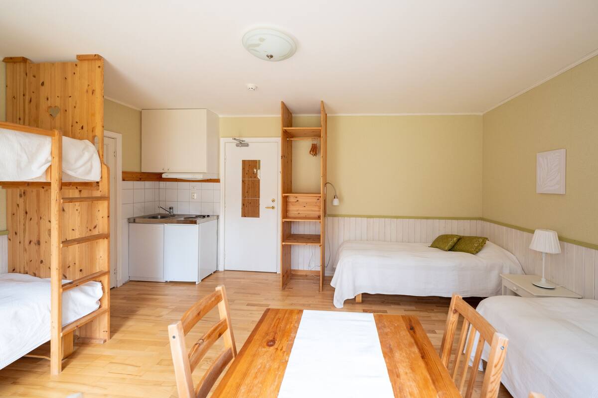 A room with bunk beds, a wooden table, a kitchen area, and a bedside table with a lamp.
