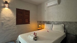 Quarto com cama, ar condicionado fixado na parede, porta de madeira e mesa de cabeceira com abajur.