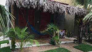 Uma casa com telhado de palha, uma rede e um pequeno jardim com palmeiras.