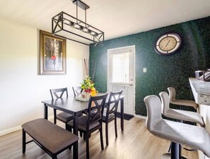 Uma sala de jantar com uma mesa de madeira, cadeiras e um banco. Há um relógio na parede e uma porta com janela.