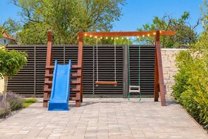 Un parque infantil con tobogán y columpios, frente a un edificio de pared con listones y vegetación.