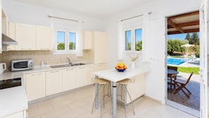 Una cocina con microondas, lavamanos y un tazón de fruta sobre la encimera. Se ve una ventana con vista a una piscina y un área de estar al aire libre.