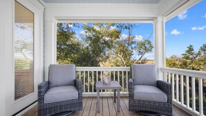 Varanda coberta com duas cadeiras de vime e uma pequena mesa, com vista para um jardim e árvores.