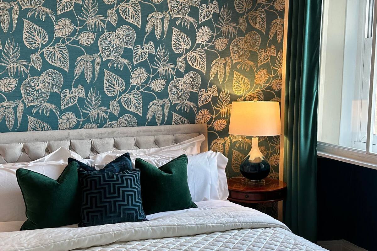 Un lit bien fait, avec une couette blanche matelassée, une tête de lit capitonnée et plusieurs oreillers, dont un de couleur verte. Une lampe est posée sur une table de chevet en bois, à côté du lit, et une fenêtre avec des rideaux verts est visible.