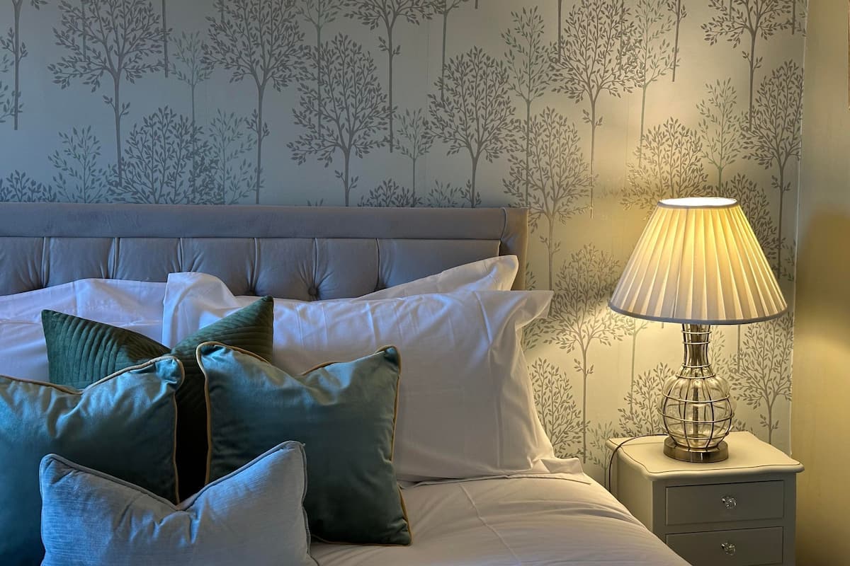Un lit bien fait, avec des oreillers blancs et verts, une tête de lit grise et une table de chevet surmontée d’une lampe.