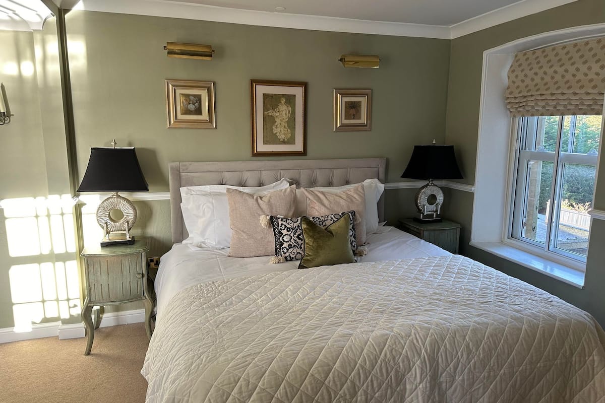 Une chambre avec un grand lit, deux tables de chevet, une fenêtre donnant sur un beau paysage et trois tableaux encadrés accrochés au mur.