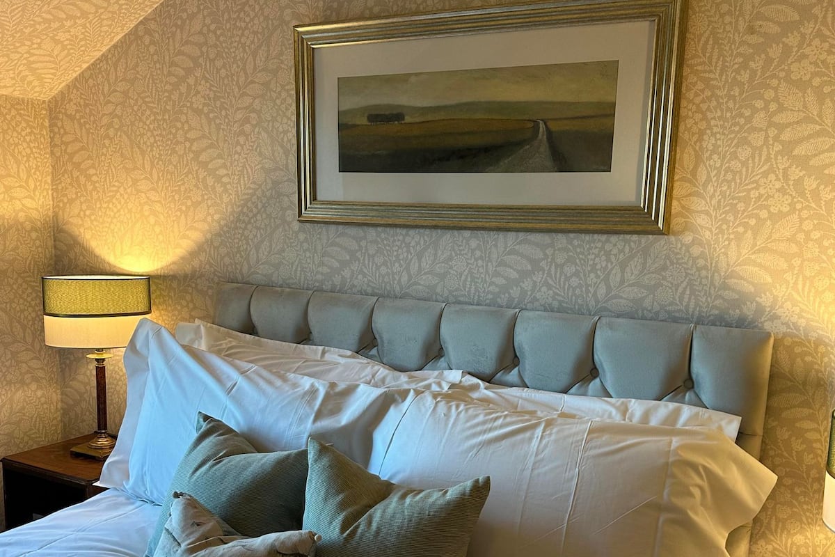 Un lit bien fait, avec une tête de lit, un tableau encadré au-dessus et une lampe sur une table de chevet.