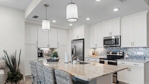 A modern kitchen with white cabinetry, stainless steel appliances, a marble island, and a dining area with wicker chairs. - Otter Lagoon at One Particular Harbour  379-103 Lagoon Views · Gaming · 3BR (Bradenton)