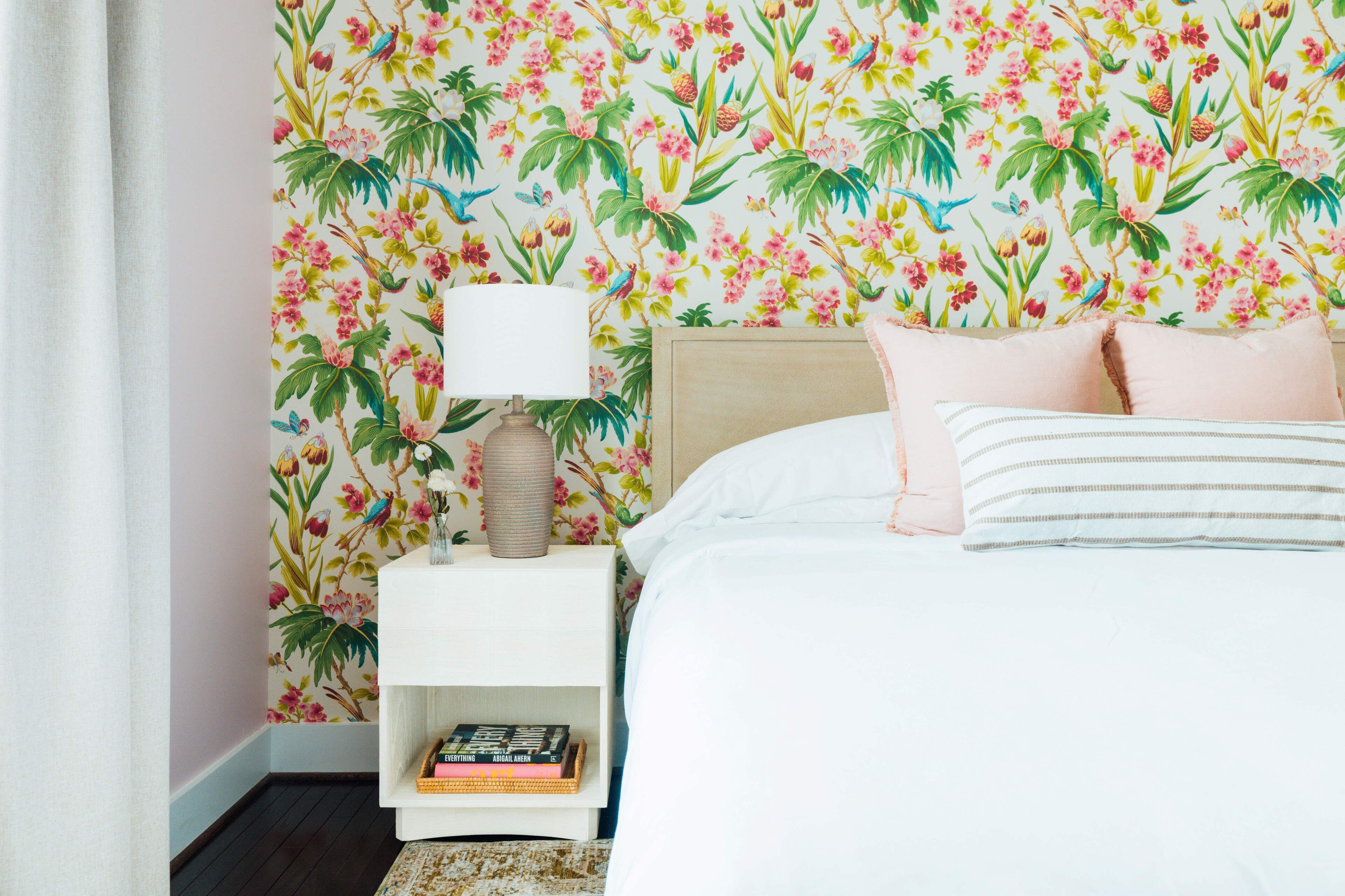 A bedroom with a floral wallpaper, a white nightstand with a lamp, a bed with pink and white bedding, and a bookshelf with books.