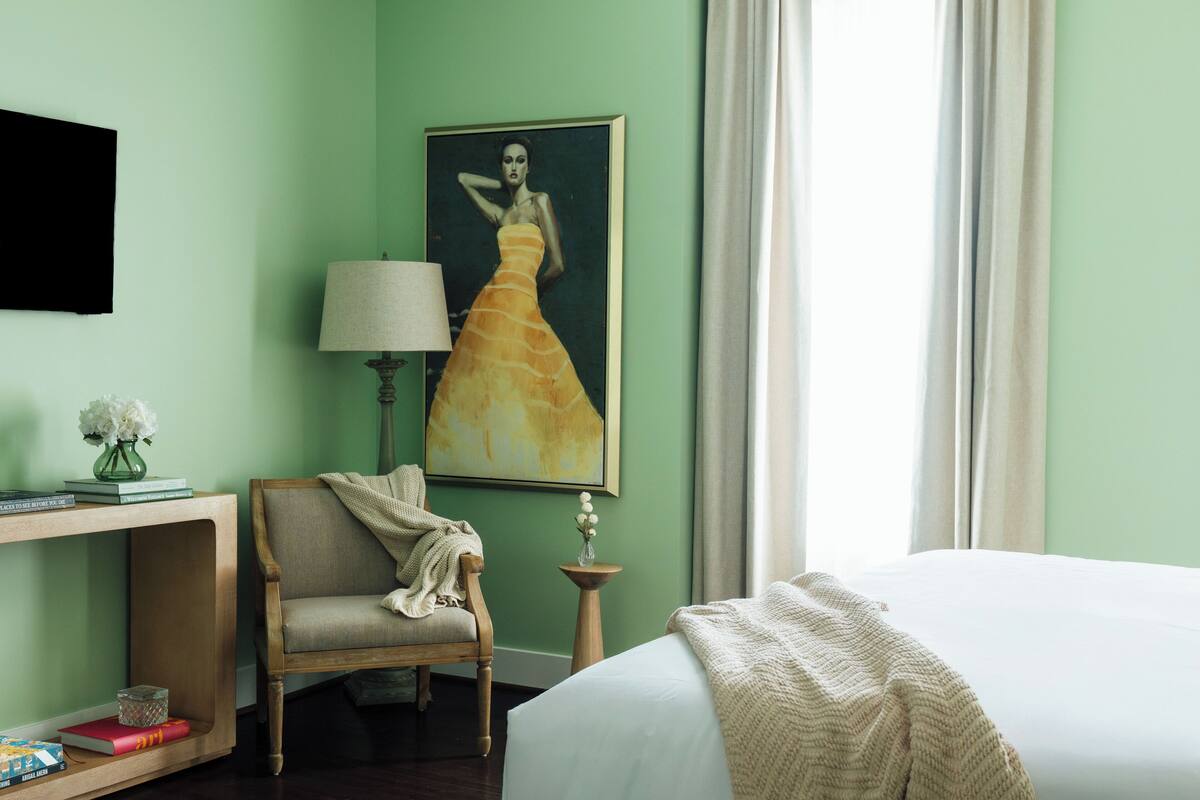 A hotel room with a bed, a chair, a lamp, a painting, and a small table with a vase.