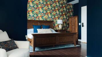 A bedroom with a wooden bed, blue and white bedding, a floral wallpaper, and a wooden nightstand with a lamp.