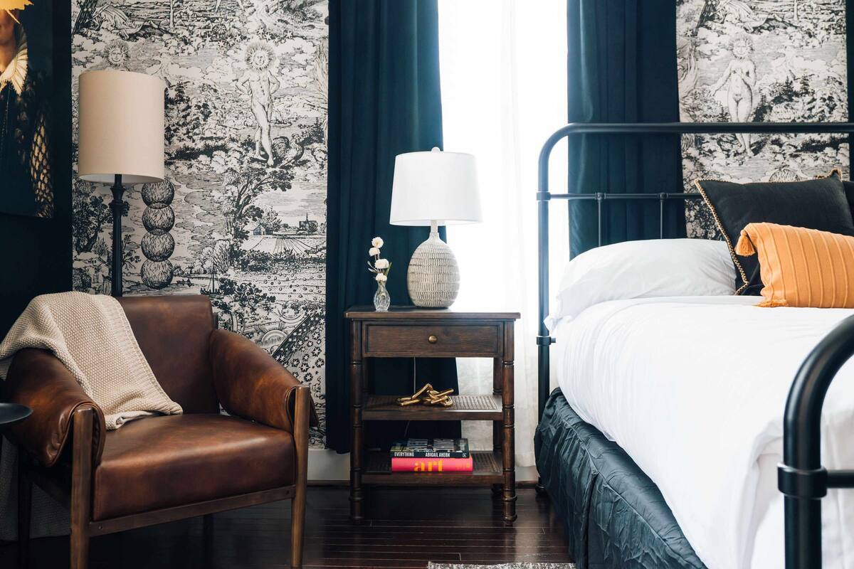 A bedroom with a bed, a nightstand with a lamp, a chair, and a bookshelf.