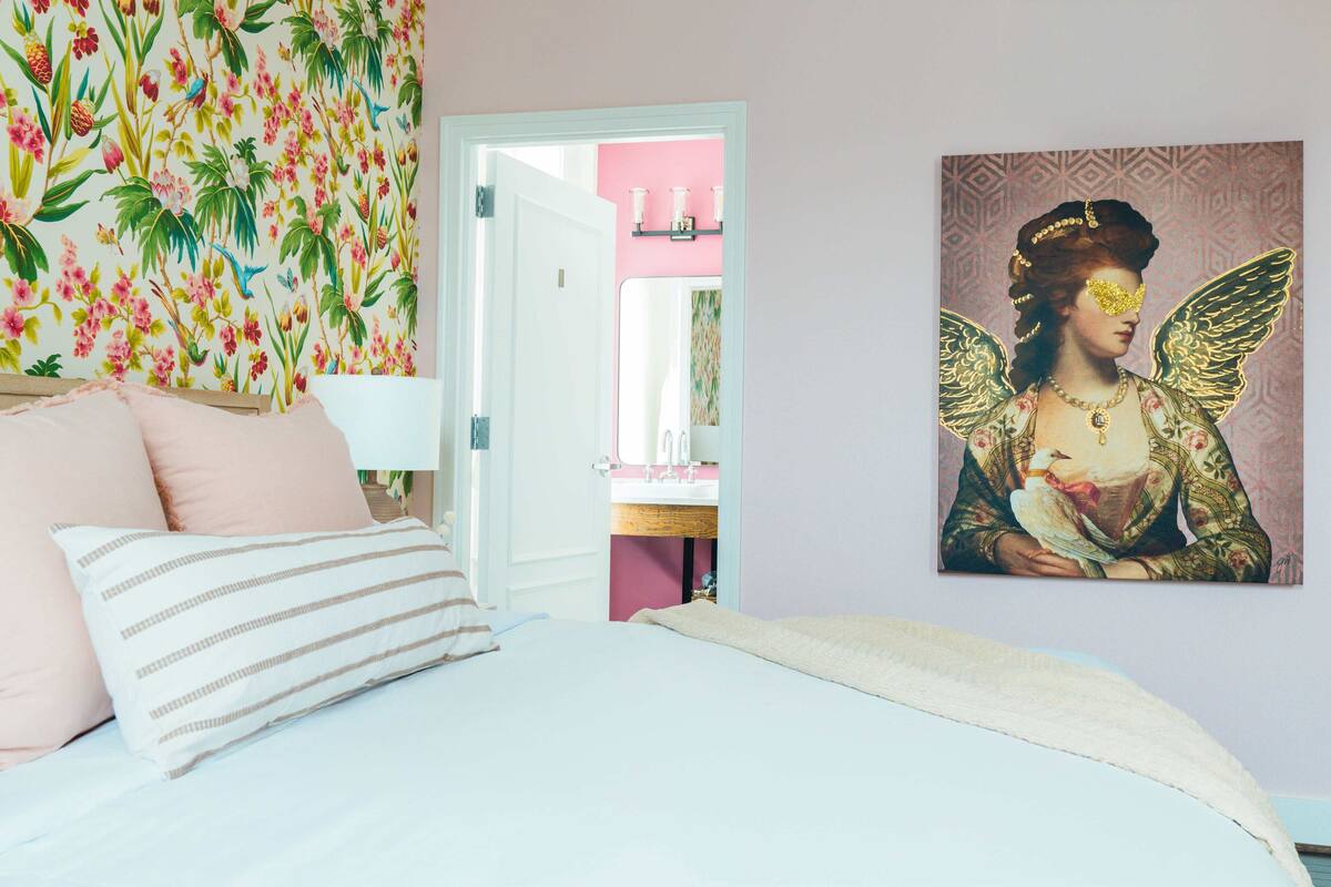 A bedroom with a bed, pillows, a wall with a floral pattern, and a painting of a figure with wings.