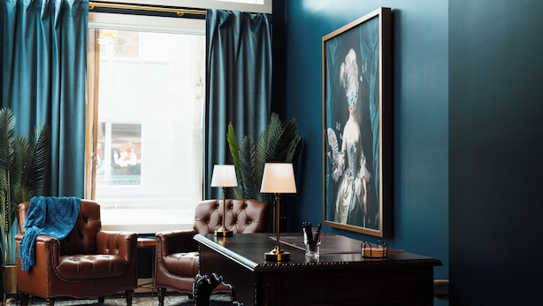 A room with a dark blue wall, a large framed painting, two leather armchairs, a wooden desk with lamps, and a window with curtains.