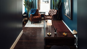 A room with a dark wooden floor, a brown leather armchair, a small table with a lamp, and a framed picture on the wall.