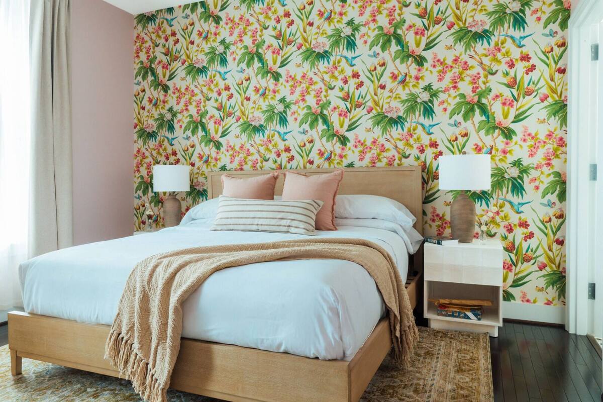 A bedroom with a floral wallpaper, a wooden bed, two bedside tables with lamps, and a window with curtains.