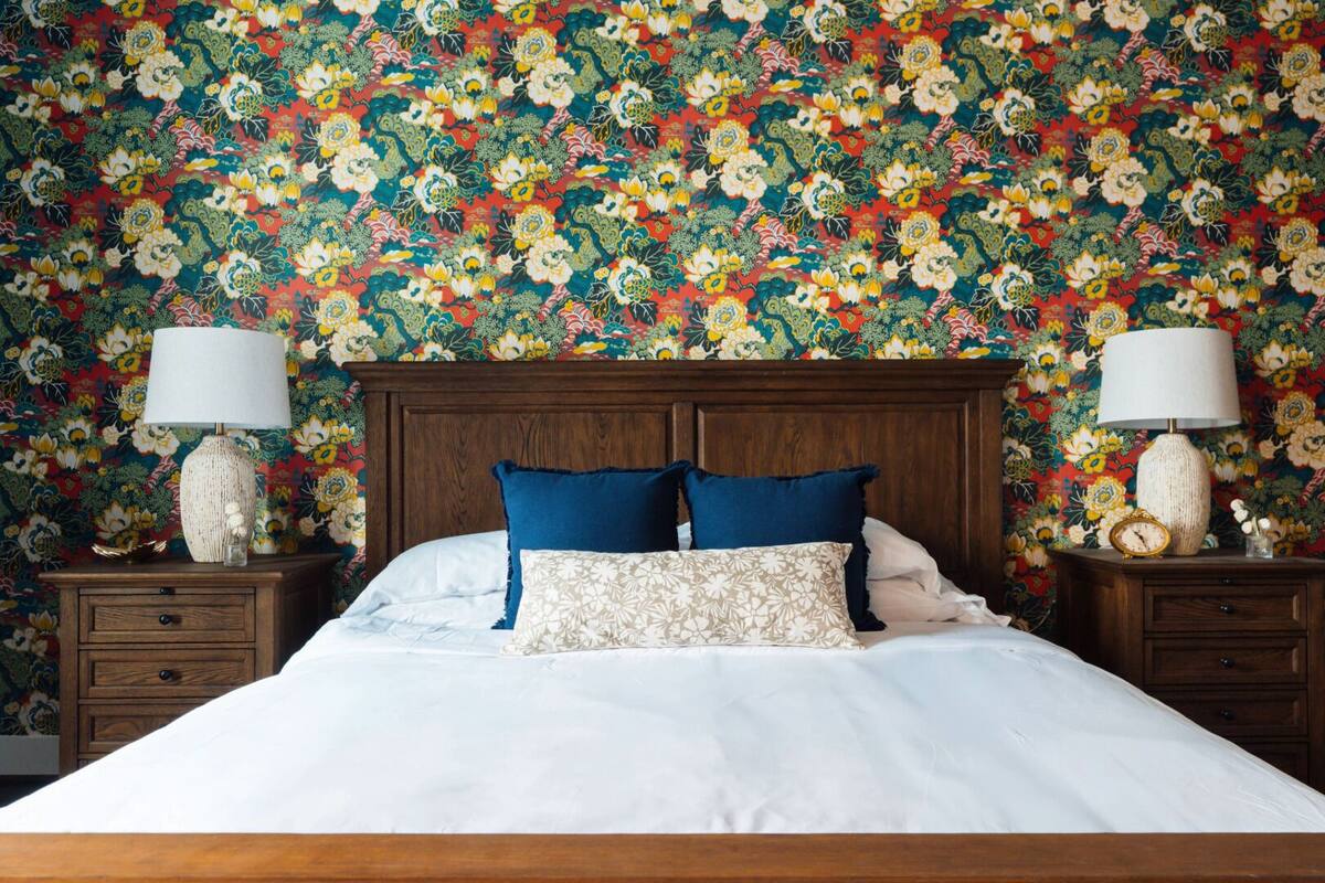 A bedroom with a floral wallpaper, a wooden headboard, two bedside tables with lamps, and a bed with white bedding and blue pillows.