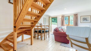 Een houten trap leidt naar een verdieping, met een eetgedeelte, een tafel, stoelen, een bank en een raam met gordijnen.