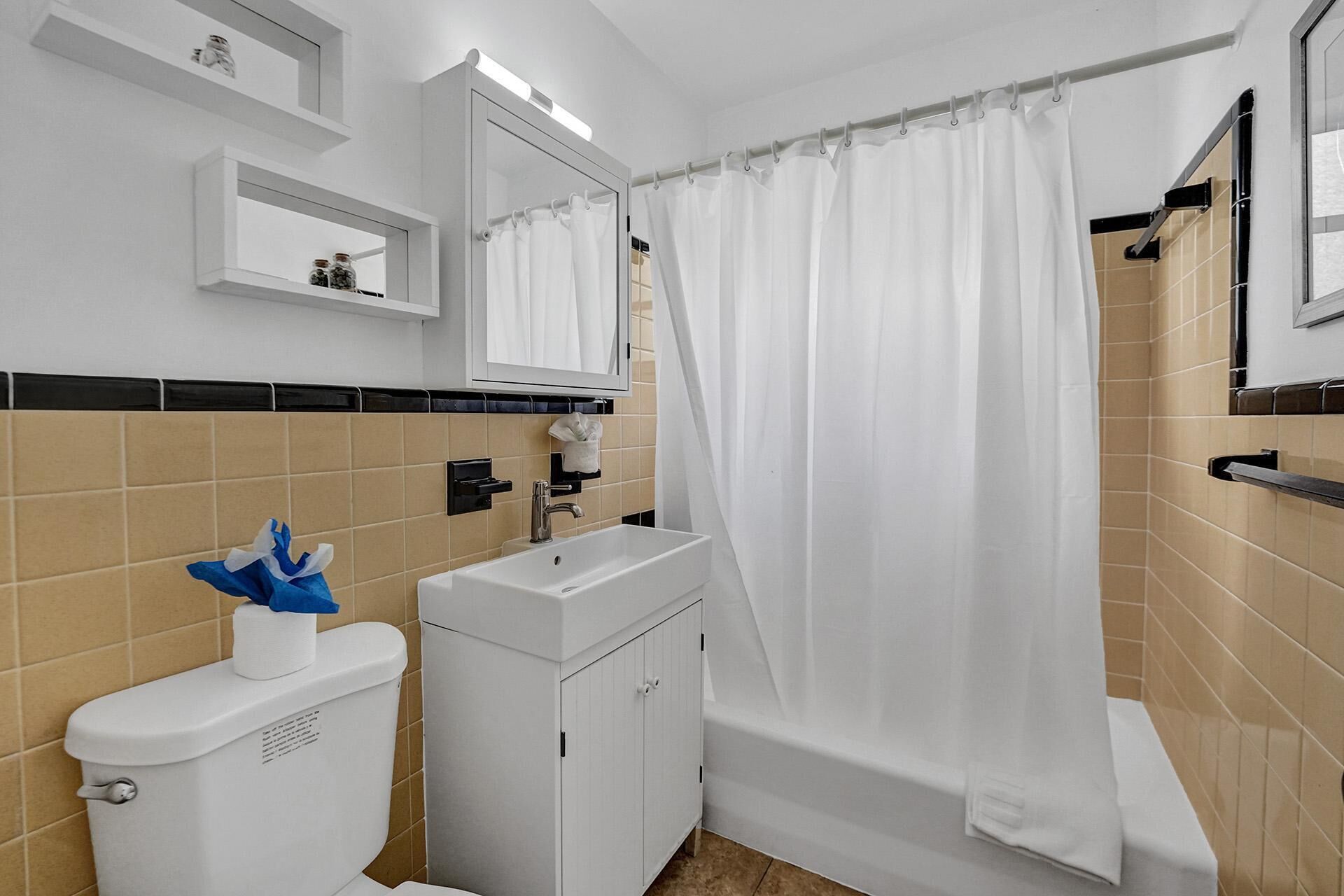 Une salle de bain avec des toilettes, un lavabo et une baignoire, tous de couleur blanche. On trouve un porte-serviettes au-dessus du lavabo et un miroir au-dessus des toilettes. Les murs sont recouverts de carreaux de couleur beige et blanche.