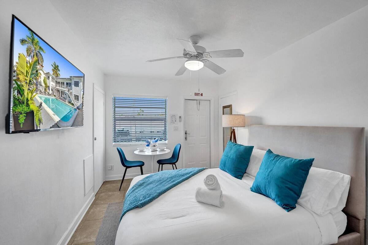Une chambre d’hôtel avec un lit, un ventilateur de plafond, un bureau avec des chaises, une fenêtre avec des stores et un téléviseur fixé au mur.