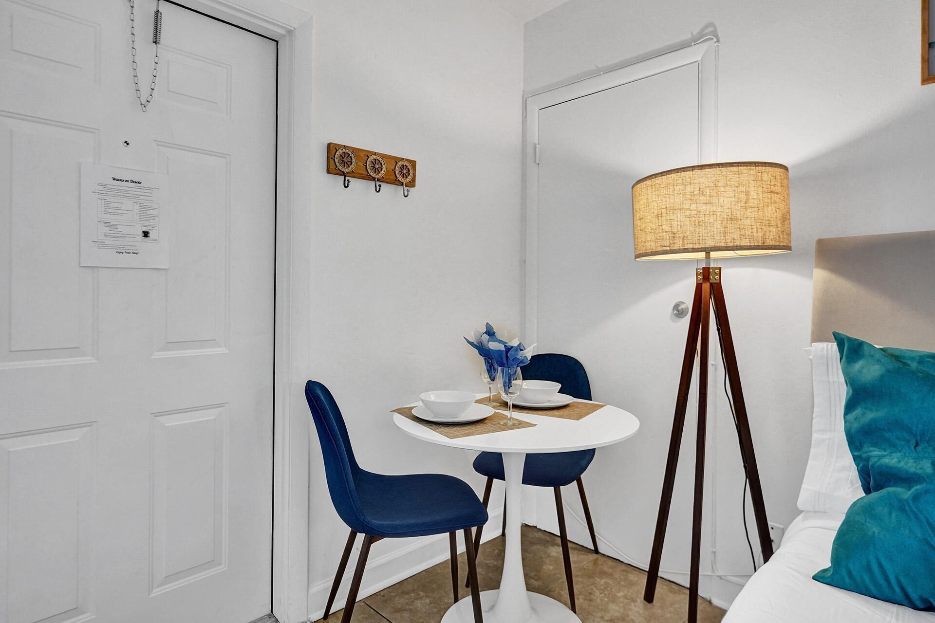 A small, neatly arranged room with a white door, a table with two chairs, a floor lamp, and a bed with teal pillows.