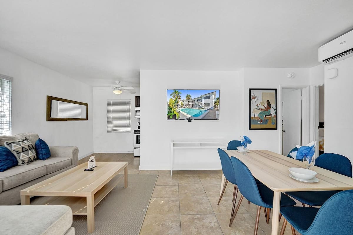 Un salon moderne comprenant un canapé, une table basse, une table à manger avec des chaises, un téléviseur fixé au mur et un coin cuisine avec une cuisinière et un four.