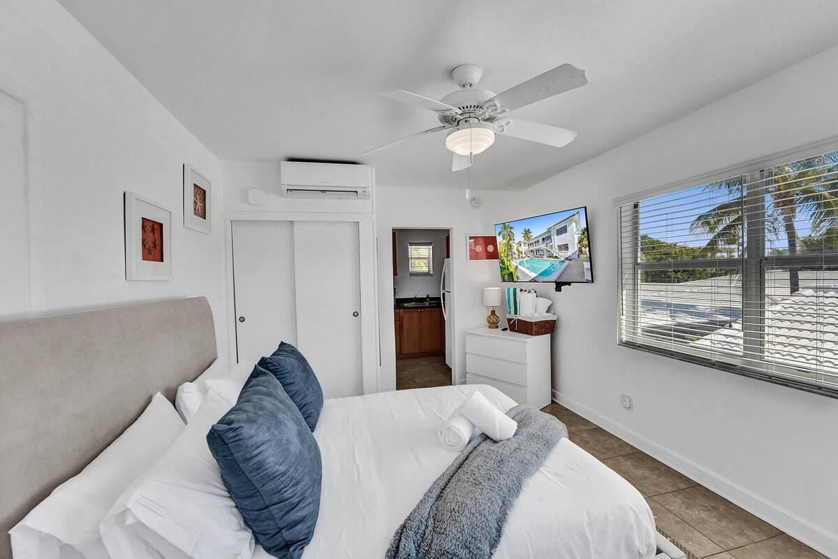 Une chambre à coucher avec un lit, un ventilateur de plafond, une télévision et une fenêtre avec des stores.