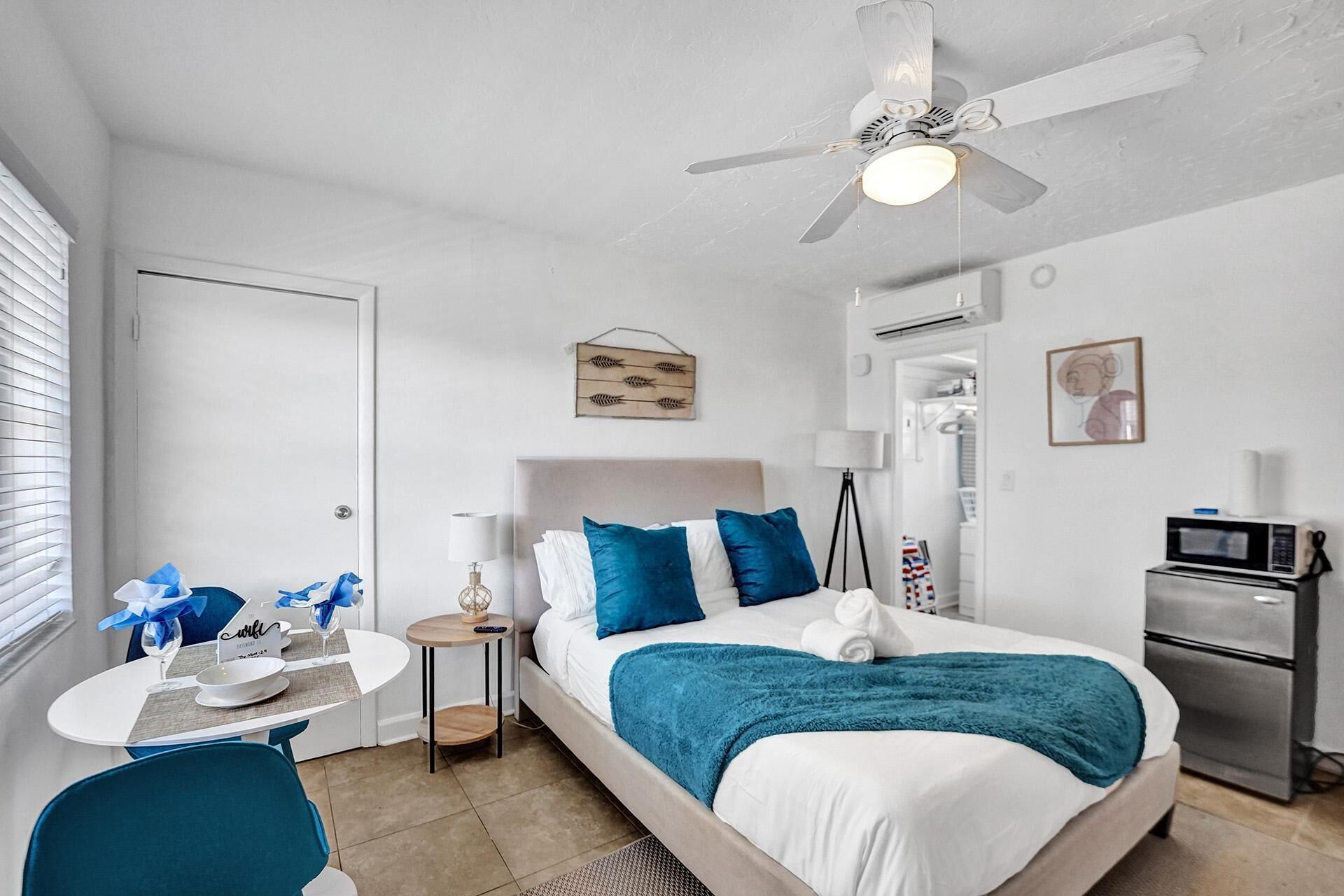 A hotel room with a bed, a desk with a chair, a small table, a ceiling fan, a microwave, and a refrigerator.