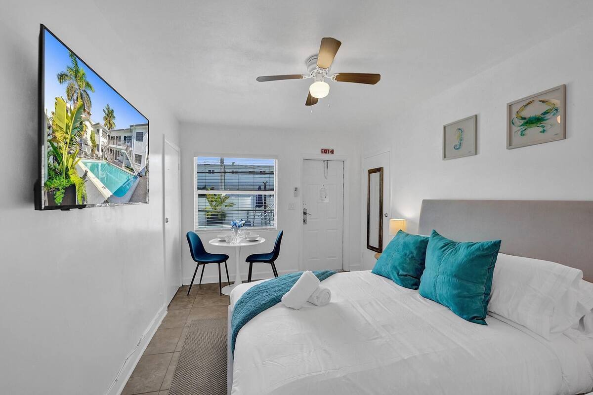 Une chambre d’hôtel avec un lit, un ventilateur de plafond, une fenêtre, une petite table avec des chaises, et une vue sur une piscine et des palmiers.