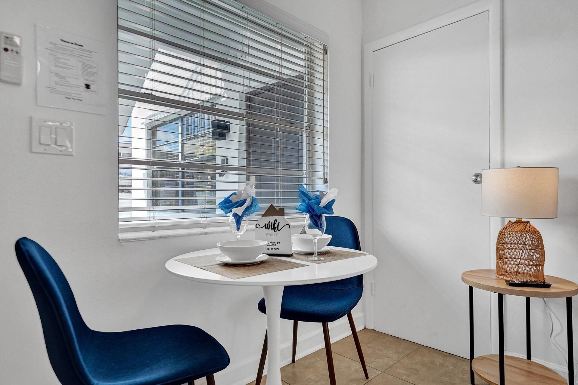 A small dining area with a round table, two blue chairs, a white lamp, and a window with blinds.