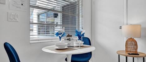 A small dining area with a round table, two blue chairs, a white lamp, and a window with blinds.