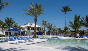 A pool area with lounge chairs and umbrellas, surrounded by palm trees and a building with a white facade. - Seaside Solace: 1BR/1Bath Condo (Fort Pierce)